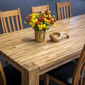 Puede incluir: Una mesa de comedor de madera clara, rodeada de sillas a juego. Un jarrón con flores amarillas y rojas está sobre la mesa, junto con un cuenco de madera. La mesa tiene forma rectangular y es de madera.
