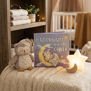 May include: A plush owl toy wearing a nightcap sits next to a children's book titled "Goodnight Dear Oliver." A star-shaped night light and a wooden crescent moon are also on display. The scene is set on a cream-colored blanket.