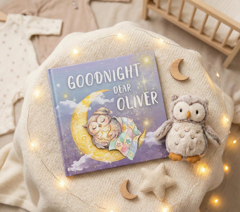 May include: A children's book titled "Goodnight Dear Oliver" with an illustration of a sleeping owl on a crescent moon. A plush owl toy, wooden moons, and a star are arranged on a cream-colored blanket, with fairy lights.