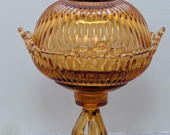 May include: Amber-colored, vintage glass candy dish with a lid. The dish has a ribbed, spherical body and sits on a pedestal base with a decorative, scalloped edge. The lid features a small handle.