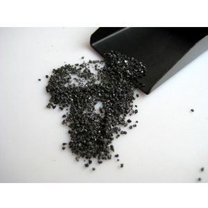 May include: A small black scoop with a pile of black sand or gravel spilling out of it onto a white surface.