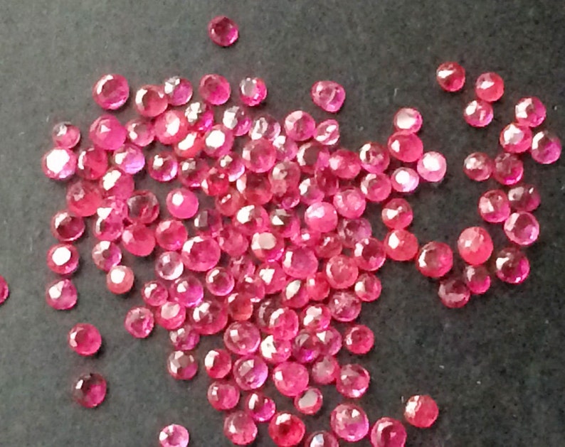 May include: A collection of faceted, round, bright red gemstones scattered across a dark surface. The gemstones vary slightly in size and reflect light, showcasing their cut and color. These appear to be rubies, suitable for jewelry making or collecting.