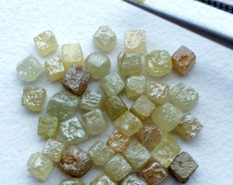 Puede incluir: Colección de diamantes en bruto, sin tallar, en varios tonos de amarillo, marrón y verde. Un diamante es sujetado por unas pinzas. Los diamantes tienen un aspecto natural, sin pulir, con formas irregulares y superficies texturizadas.