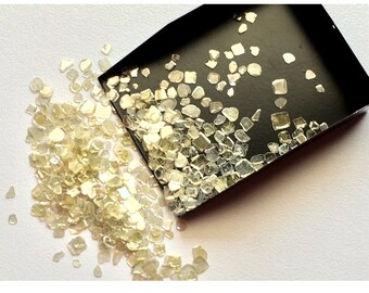 May include: A small black tray filled with loose, iridescent, light yellow, and clear diamond chips. The tray is tipped over, spilling the chips onto a white surface.