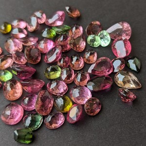 May include: A collection of faceted gemstones in various shapes and sizes, including oval, pear, and round cuts. The gems display a range of colors, from pink and purple to green and yellow, with a polished, reflective surface. The gemstones are scattered on a dark surface.