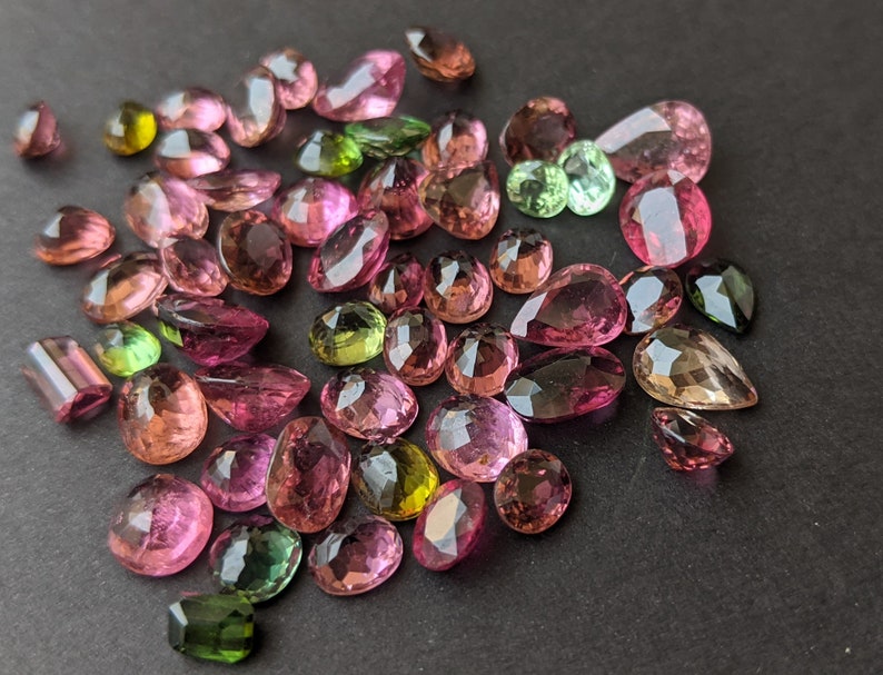 May include: A collection of faceted gemstones in various shapes and sizes, including oval, pear, and round cuts. The gems display a range of colors, from pink and purple to green and yellow, with a polished, reflective surface. The gemstones are scattered on a dark surface.