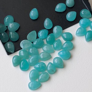 May include: A collection of loose, teardrop-shaped turquoise gemstones with a smooth, polished finish. The stones exhibit a vibrant, semi-translucent blue-green hue and are scattered across a black tray and a plain white surface.