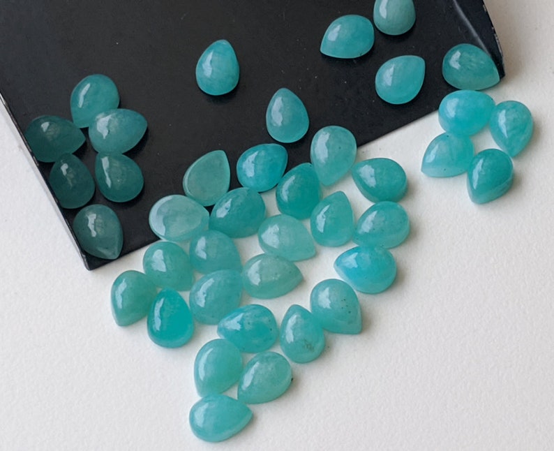 May include: A collection of loose, teardrop-shaped turquoise gemstones with a smooth, polished finish. The stones exhibit a vibrant, semi-translucent blue-green hue and are scattered across a black tray and a plain white surface.