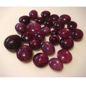 May include: A collection of 20 star ruby cabochons in various sizes and shades of purple. The cabochons are displayed on a white surface.