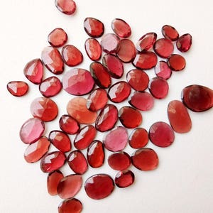 May include: A collection of faceted, oval and rounded gemstones in shades of red and pink. The gemstones are scattered on a white surface, with varying sizes and shapes, showcasing their polished surfaces.