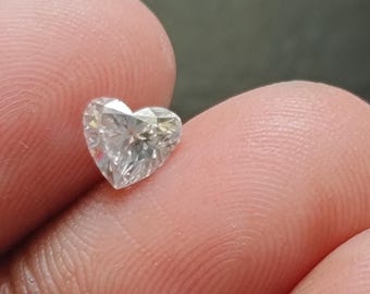 Può includere: Una piccola pietra preziosa trasparente a forma di cuore con molteplici sfaccettature che riflettono la luce. La pietra è incolore e traslucida, caratterizzata da un taglio brillante con bordi netti e una base appuntita.