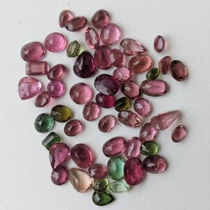 May include: A collection of faceted gemstones in various shapes and sizes. The gems are primarily pink and purple, with a few green stones interspersed. The gemstones are scattered on a white surface.