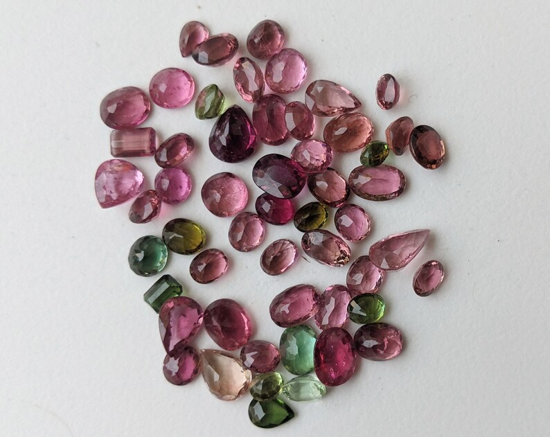 May include: A collection of faceted gemstones in various shapes and sizes. The gems are primarily pink and purple, with a few green stones interspersed. The gemstones are scattered on a white surface.