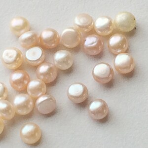 May include: A collection of approximately 20 small, round, off-white pearls with a slight pink hue. The pearls are smooth and have a glossy finish.