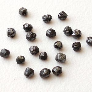 May include: A collection of black diamonds, also known as carbonado diamonds, on a white surface. These rough diamonds are naturally occurring and have a dark, opaque appearance.
