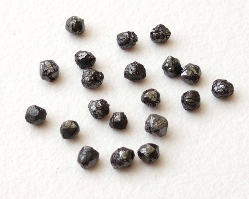 May include: A collection of black diamonds, also known as carbonado diamonds, on a white surface. These rough diamonds are naturally occurring and have a dark, opaque appearance.