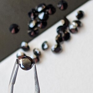 May include: A single black gemstone held in tweezers. The gemstone is round and faceted. There are other black gemstones scattered on a white surface.