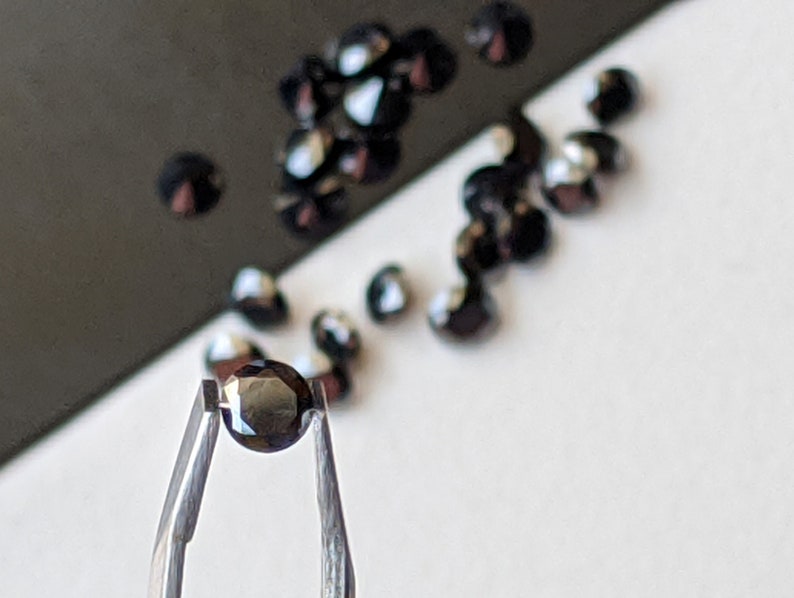 May include: A single black gemstone held in tweezers. The gemstone is round and faceted. There are other black gemstones scattered on a white surface.