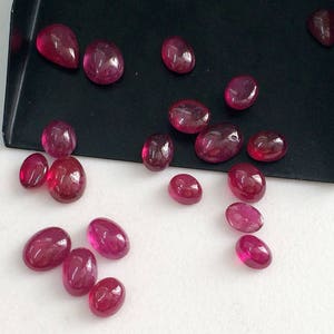 May include: A collection of oval-shaped, faceted, ruby red gemstones. The gemstones are scattered on a white surface.