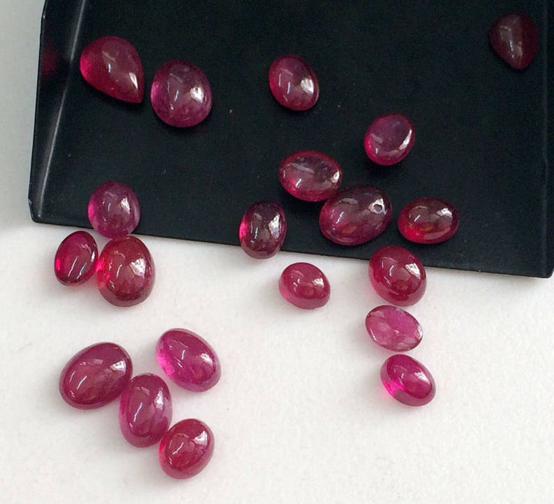 May include: A collection of oval-shaped, faceted, ruby red gemstones. The gemstones are scattered on a white surface.