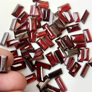 May include: A collection of red rectangular gemstones, likely garnets, scattered on a white surface. The gemstones are faceted and have a glossy finish.