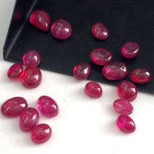 May include: A collection of 16 oval and teardrop shaped ruby gemstones. The gemstones are various shades of red and are arranged on a white surface.
