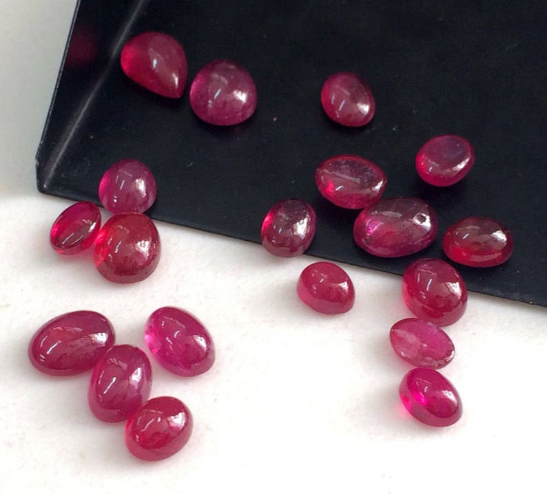 May include: A collection of 16 oval and teardrop shaped ruby gemstones. The gemstones are various shades of red and are arranged on a white surface.