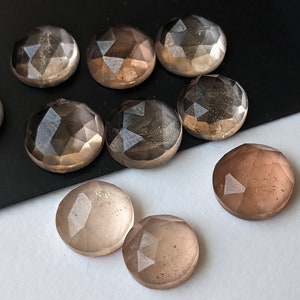 Puede incluir: Una colecci&oacute;n de ocho cabujones de piedra preciosa redondos y facetados, transl&uacute;cidos, en tonos de marr&oacute;n y gris. Los cabujones est&aacute;n dispuestos sobre una superficie negra.