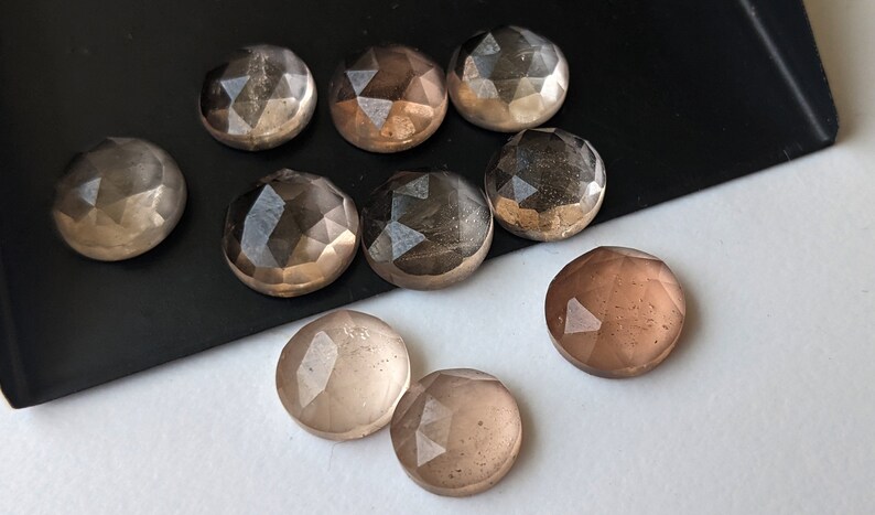 Puede incluir: Una colecci&oacute;n de ocho cabujones de piedra preciosa redondos y facetados, transl&uacute;cidos, en tonos de marr&oacute;n y gris. Los cabujones est&aacute;n dispuestos sobre una superficie negra.