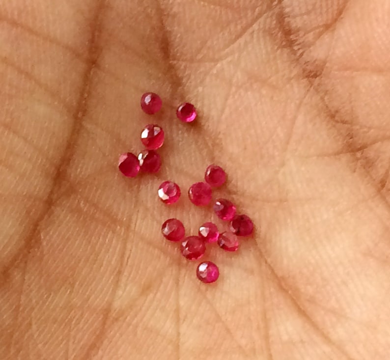 May include: A collection of small, faceted, round, ruby gemstones. The gems are a vibrant red color and are scattered across a light brown surface. The gemstones are likely used for jewelry making or other crafts.