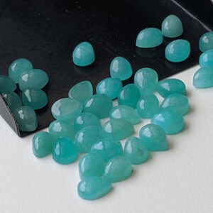May include: A collection of small, smooth, teardrop-shaped turquoise gemstones with a polished finish. The stones are scattered across a surface, with several resting inside a black metal scoop, displaying a consistent vibrant blue-green color.