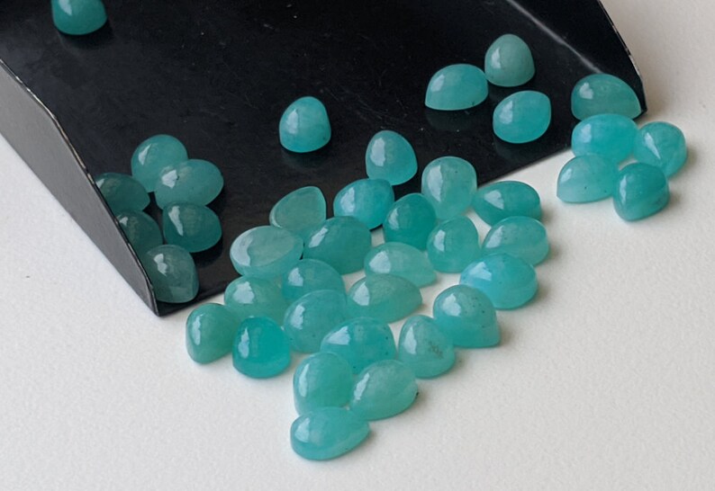 May include: A collection of small, smooth, teardrop-shaped turquoise gemstones with a polished finish. The stones are scattered across a surface, with several resting inside a black metal scoop, displaying a consistent vibrant blue-green color.