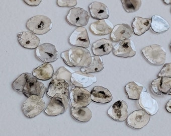 May include: A collection of small, rough, gray diamonds, also known as rough diamonds, scattered on a white surface.