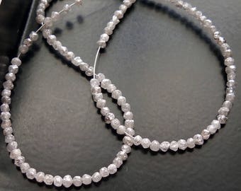May include: Two strands of raw diamond beads, strung on a thin, white cord. The beads are a light gray color and have a faceted, irregular shape. The strands are arranged in a looping pattern against a dark background.
