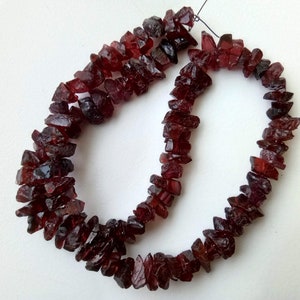Puede incluir: Una hebra de cuentas de piedras preciosas de color rojo oscuro, cada cuenta tiene una forma irregular y tosca. Las cuentas están enhebradas en un cordón negro delgado.