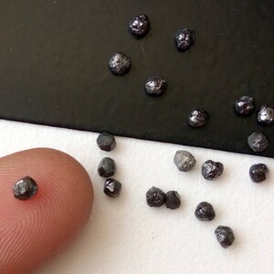 May include: A collection of small, black, rough diamonds on a white surface. Some of the diamonds are on a finger for size comparison.