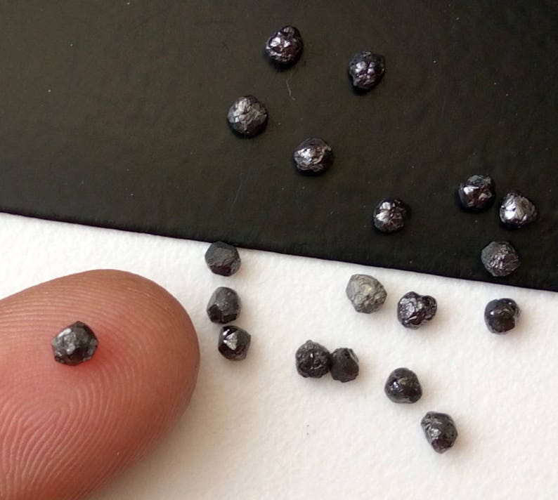 May include: A collection of small, black, rough diamonds on a white surface. Some of the diamonds are on a finger for size comparison.
