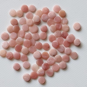 May include: A large group of pink, round, flat, smooth, polished stones. The stones are scattered on a white surface.
