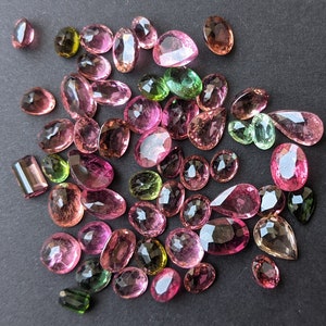 May include: A collection of faceted gemstones in various shapes and sizes, including oval, pear, and rectangular cuts. The gems display a range of colors, from pink and rose to green and brown, with a sparkling, polished finish. The gemstones are scattered on a dark surface.