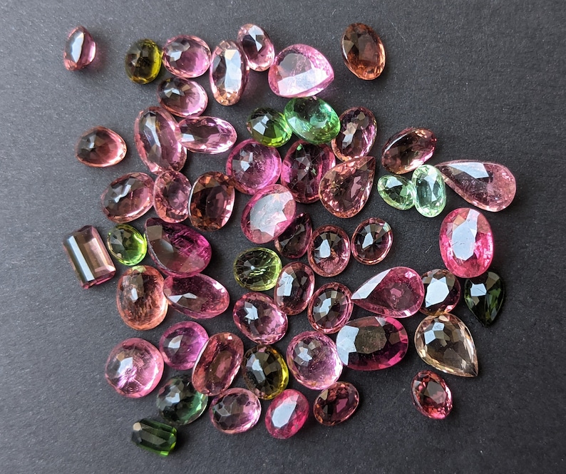 May include: A collection of faceted gemstones in various shapes and sizes, including oval, pear, and rectangular cuts. The gems display a range of colors, from pink and rose to green and brown, with a sparkling, polished finish. The gemstones are scattered on a dark surface.