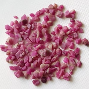 Könnte beinhalten: Eine Ansammlung kleiner, grob geschnittener Rubin-Edelsteine. Die Edelsteine haben eine leuchtende rosa-rote Farbe mit unterschiedlichen Formen und Größen. Sie sind auf einer weißen Oberfläche verstreut und erzeugen ein strukturiertes Aussehen. Die Rohrubinkristalle sind ideal für die Schmuckherstellung.