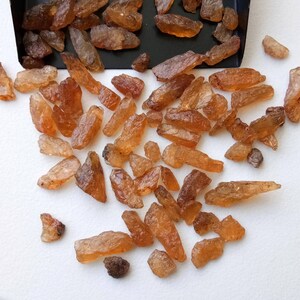 May include: A collection of raw, rough, orange-brown gemstones. The stones are various sizes and shapes, and some have a slightly translucent quality.