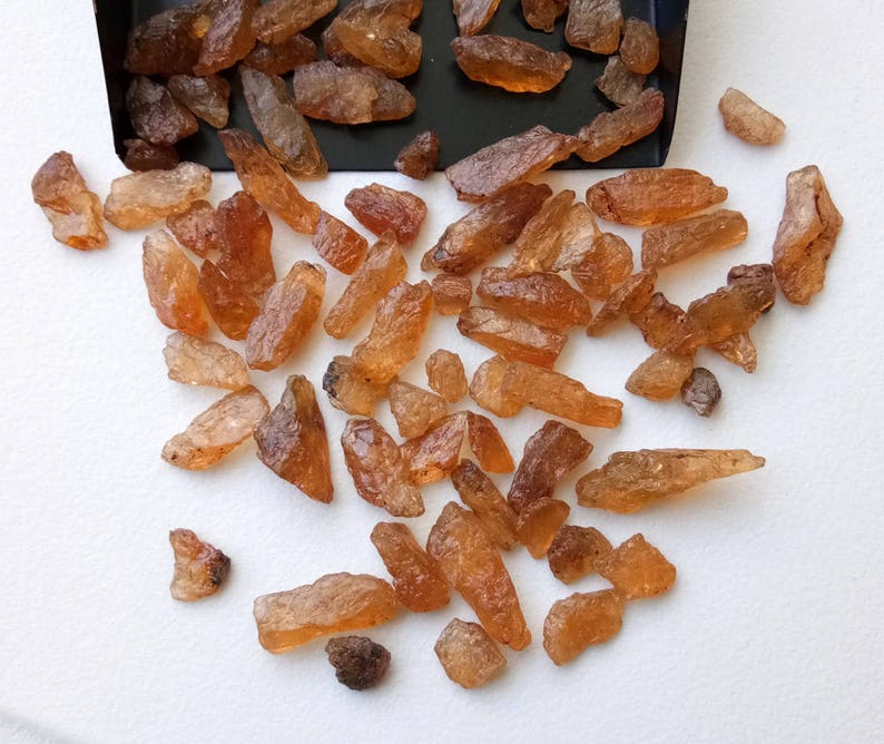 May include: A collection of raw, rough, orange-brown gemstones. The stones are various sizes and shapes, and some have a slightly translucent quality.