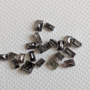 May include: A collection of small, black, rectangular, faceted gemstones. The stones have a rough, natural finish and are scattered on a white surface.