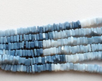 Puede incluir: Una hebra de cuentas de piedras preciosas azules y blancas. Las cuentas son rectangulares y tienen una forma ligeramente irregular. Las cuentas están dispuestas en un degradado de azul oscuro a blanco.