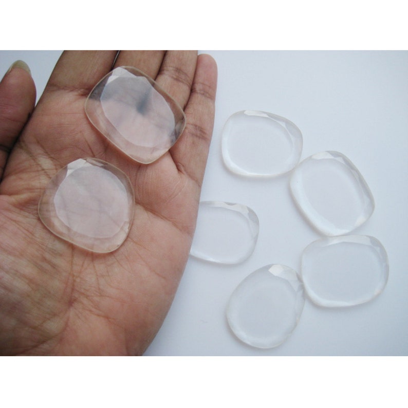 May include: Seven clear, smooth, oval-shaped crystal stones on a white surface. The stones are various sizes and are arranged in a semi-circle.