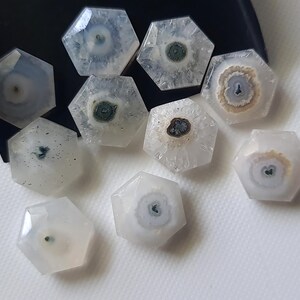 May include: A collection of nine white, hexagonal, agate cabochons with various patterns of brown, white, and green inclusions. The cabochons are arranged on a white surface.