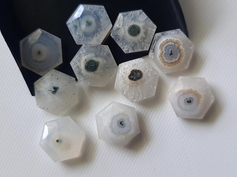 May include: A collection of nine white, hexagonal, agate cabochons with various patterns of brown, white, and green inclusions. The cabochons are arranged on a white surface.