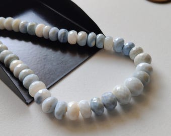 Puede incluir: Un collar o pulsera con cuentas facetadas de piedras preciosas en tonos azul pálido, blanco y crema. Las cuentas varían en tamaño y forma. La joya se presenta sobre una bandeja negra, sobre un fondo blanco.