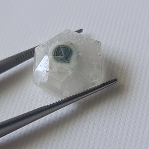 May include: A clear, faceted, hexagonal crystal with a blue and white inclusion in the center. The crystal is held by tweezers.